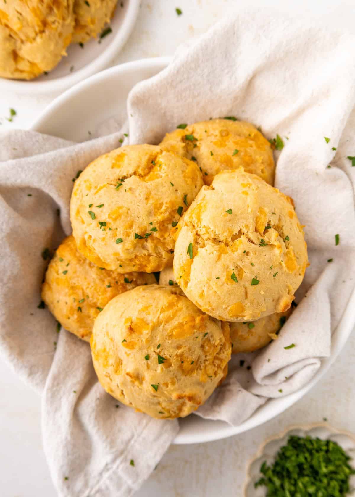 Six golden biscuits topped with chopped herbs in a bowl lined with a light-colored cloth.
