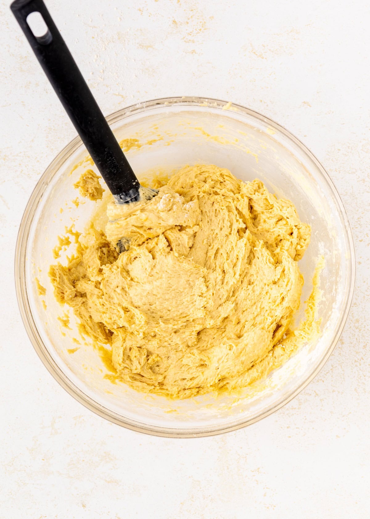 A glass bowl with yellow cookie dough and a black spatula on a light surface.