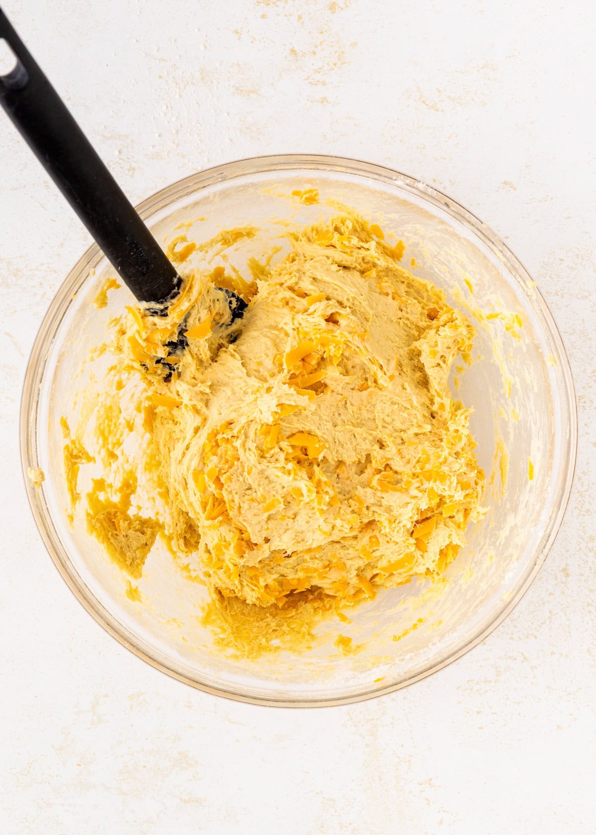 A glass bowl of cookie dough with yellow bits and a black spatula on a white surface.