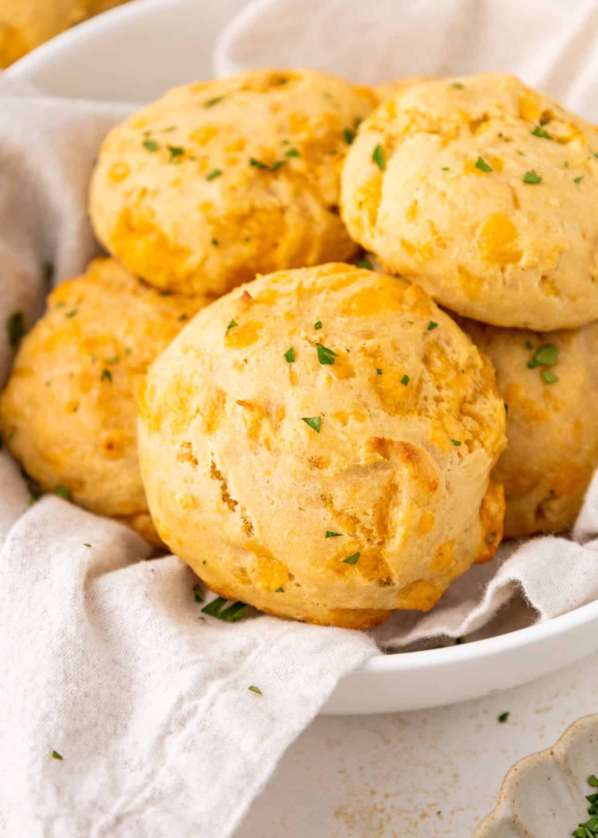 Golden cheddar biscuits topped with chopped herbs on a white cloth in a bowl.