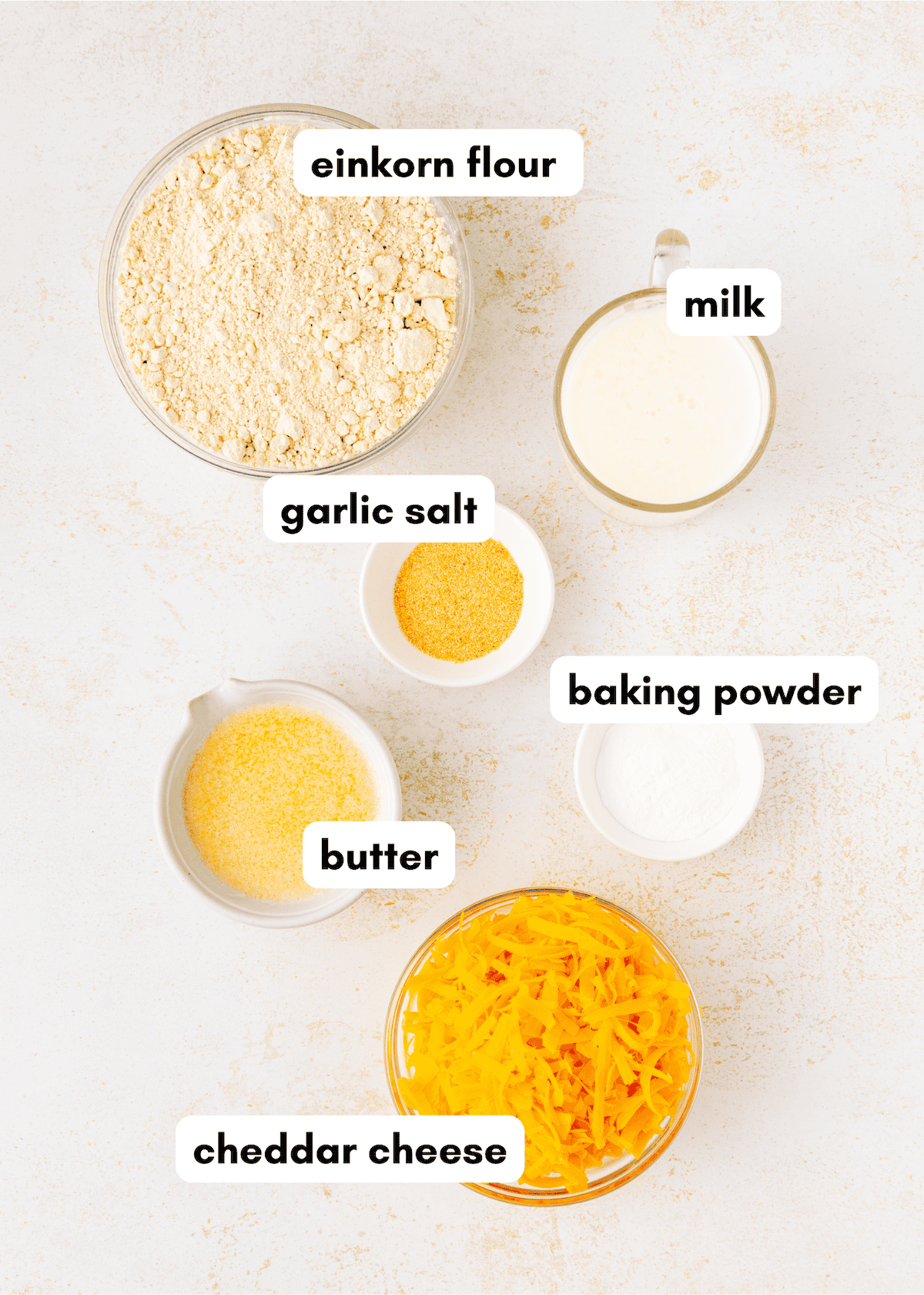 Bowls of einkorn flour, milk, garlic salt, baking powder, butter, and cheddar cheese on a table.